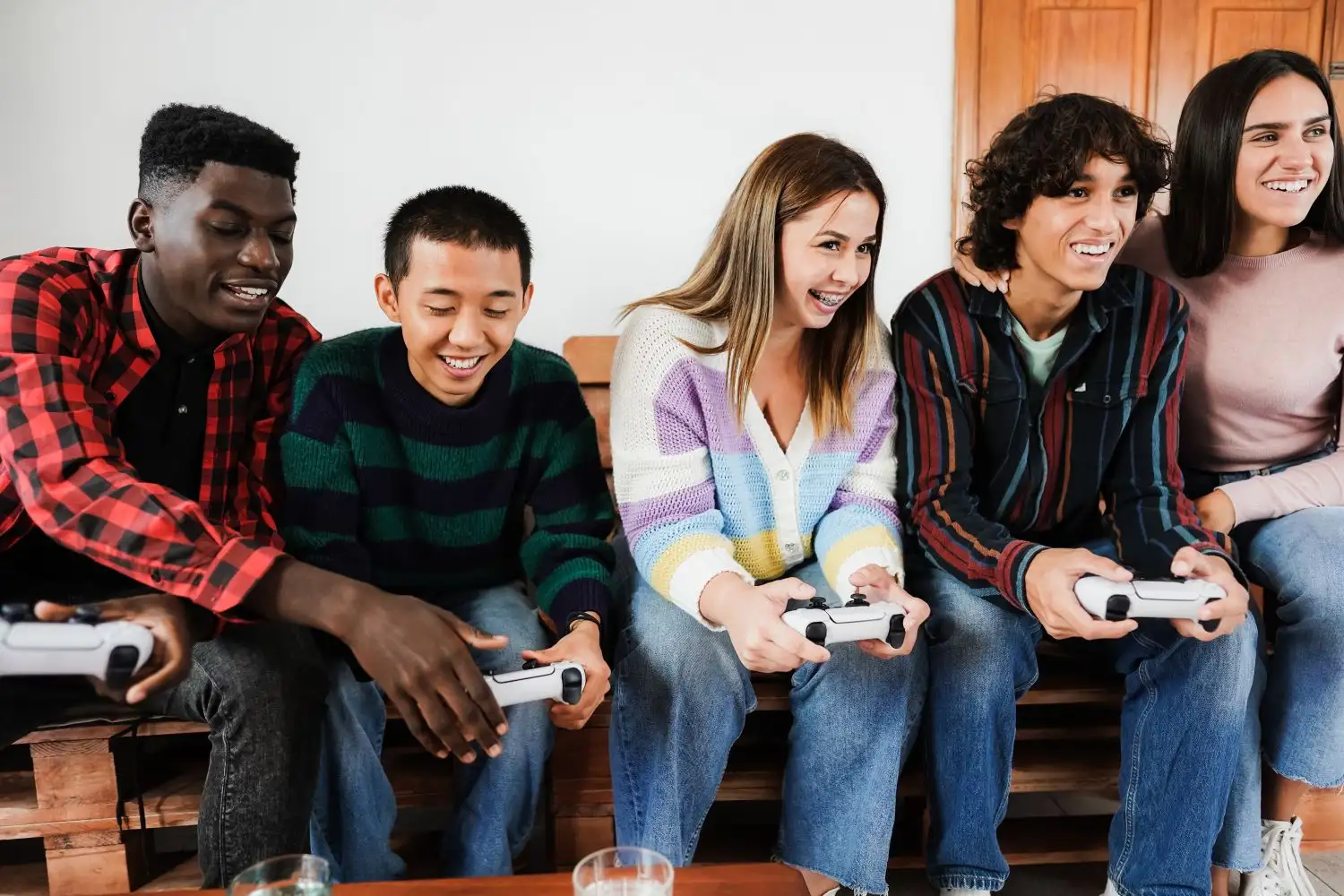 A diverse group of five young friends sitting side-by-side on a wooden pallet bench, laughing and smiling as they play a multiplayer video game together with white controllers.